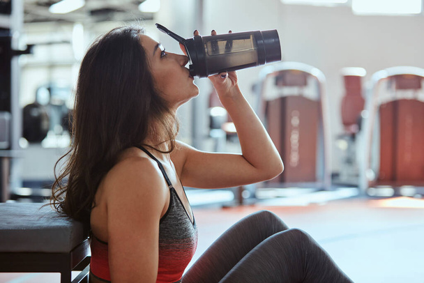 Młoda aktywna kobieta robi sobie przerwę w siłowni i pije shake 'a proteinowego. - Zdjęcie, obraz
