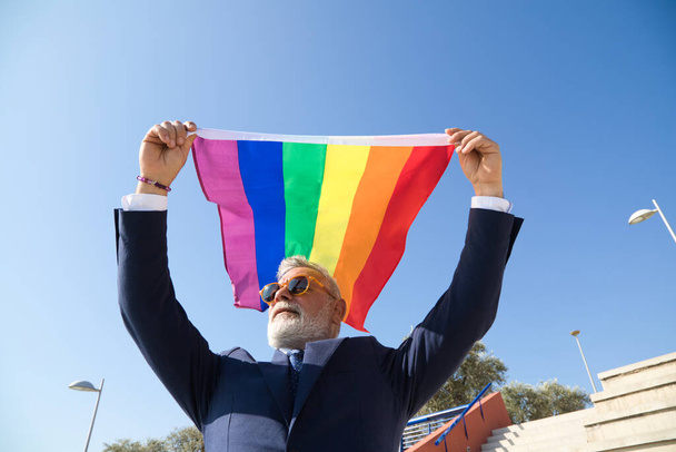 Зрелый гей, исполнительный, седой, бородатый, солнцезащитные очки, пиджак и галстук, размахивая новым флагом lgbtiq + гордости на ветру под голубым небом. Понятие зрелого гея, lgbh, гордость, равенство, права. - Фото, изображение