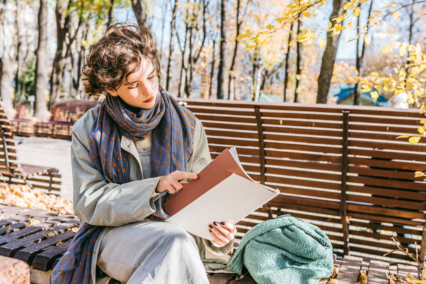 teenage girl with brown hair, autumn casual clothes, with light scarf around neck, sits alone in autumn park on bench, holds notebook and pen in hands, flips through pages, thoughtful facial expression - Photo, Image