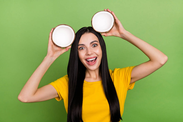 Photo of funny sweet woman wear yellow t-shirt holding head two coconut halves isolated green color background - Фото, изображение