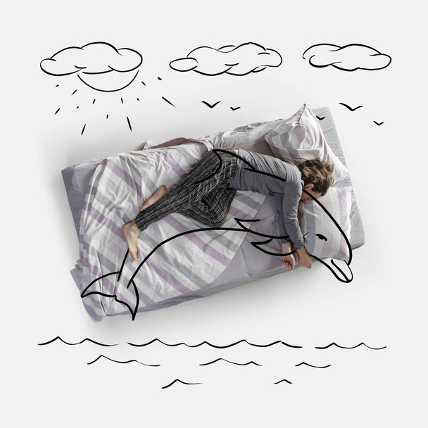 Creative image. Top view of young woman lying on bed, sleeping, dreaming of huggind dolphin - 写真・画像
