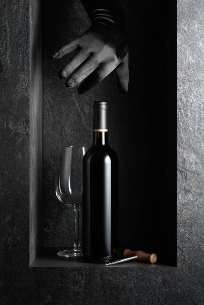 Siyah eldivenli bir kadının eli bir şişe kırmızı şaraba uzanıyor. Pahalı şarapların teması üzerine bir konsept resim. - Fotoğraf, Görsel