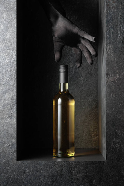 De hand van een vrouw in een zwarte handschoen reikt naar een fles witte wijn. Een concept imago op het thema van dure wijnen. - Foto, afbeelding