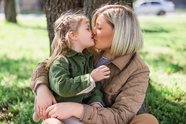 blonde woman kissing little daughter while sitting on green grass in park - Фото, изображение