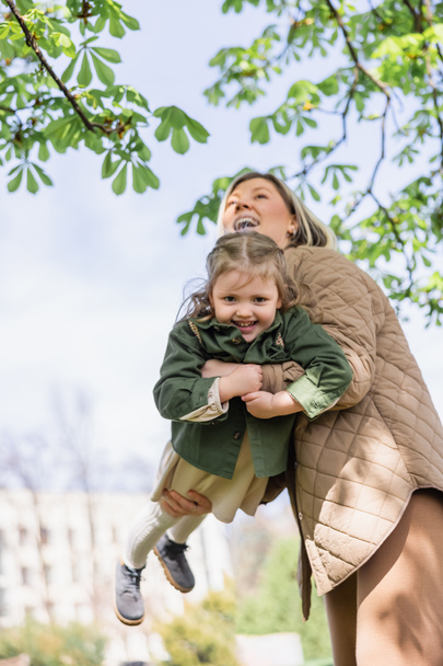 low angle view of excited girl looking at camera while mother holding her in park - Photo, image