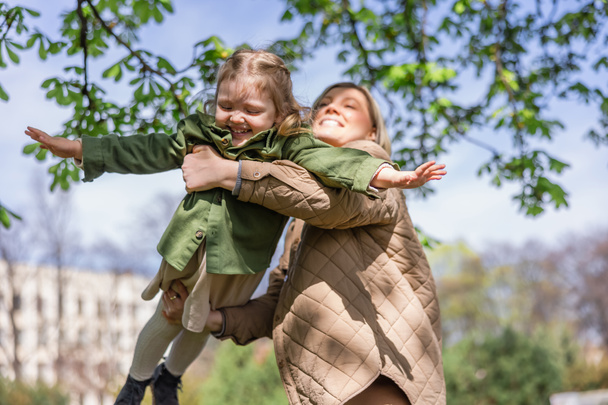 bajo ángulo vista de feliz mujer sosteniendo alegre hija al aire libre - Foto, imagen