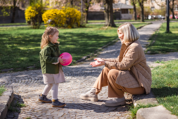boční pohled na radostnou dívku držící míč při zábavě s maminkou v parku - Fotografie, Obrázek