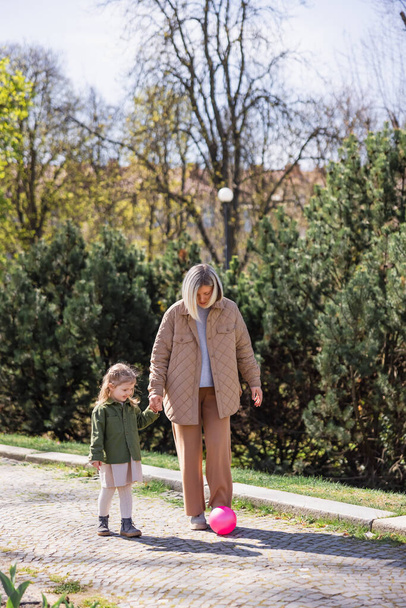 plný pohled na matku a dceru držící se za ruce při chůzi v blízkosti míče v parku - Fotografie, Obrázek