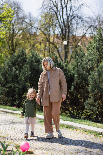 full length view of mother and child in stylish clothes holding hands and walking in park - Photo, Image