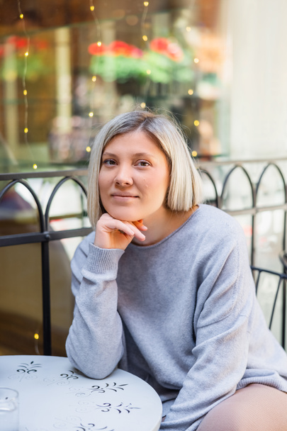 mujer rubia feliz mirando a la cámara mientras está sentado en la cafetería de la calle - Foto, Imagen