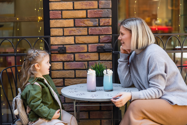 side view of excited woman sitting near daughter and glasses with milkshake in street cafe - Foto, immagini