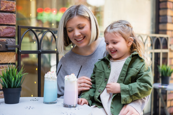 χαμογελαστό κορίτσι κοιτάζοντας νόστιμο επιδόρπιο γάλα κοντά ευτυχισμένη μητέρα στο δρόμο καφέ - Φωτογραφία, εικόνα