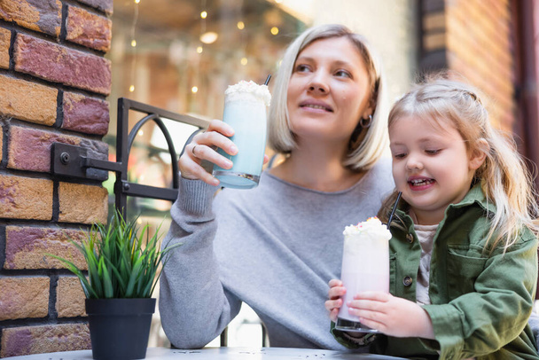 smiling girl holding glass of creamy milkshake near little daughter in street cafe - Fotoğraf, Görsel