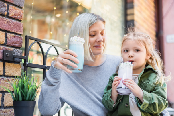 Маленькая девочка пьет вкусный молочный коктейль возле улыбающейся мамы в уличном кафе - Фото, изображение