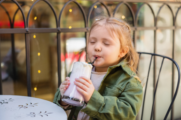 довольная девушка с закрытыми глазами пьет молочный коктейль в уличном кафе - Фото, изображение