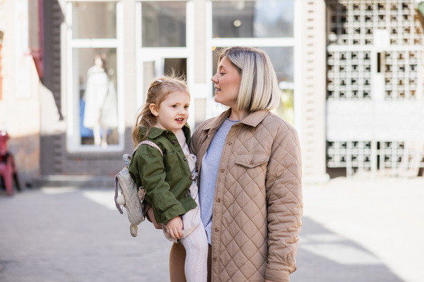 blonde woman in trendy clothes holding smiling child on city street - Foto, Imagen