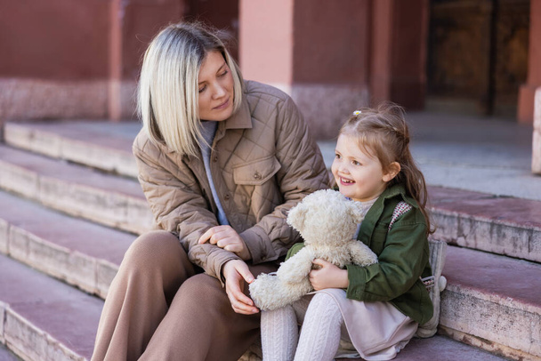 blonde woman looking at happy daughter sitting on stairs with teddy bear - Foto, Imagen