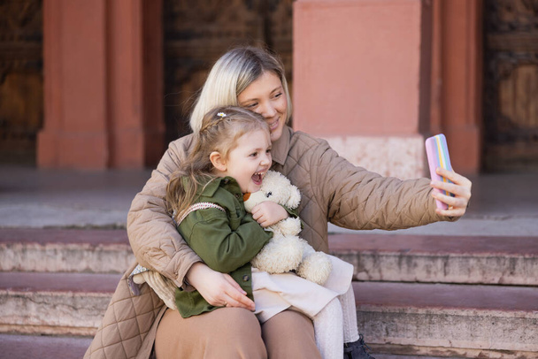 lachendes Mädchen mit Teddybär in der Nähe von Mama, die im Freien ein Selfie mit dem Smartphone macht - Foto, Bild