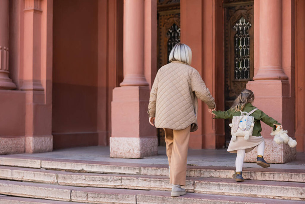 takaisin näkymä nainen ja lapsi nallekarhu kädestä kävellessään portaissa lähellä rakennusta - Valokuva, kuva
