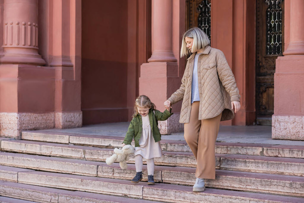 mother and daughter with teddy bear walking on stairs outdoors - Фото, изображение