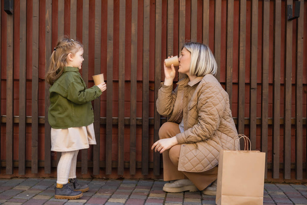 vista lateral de la madre y la hija en traje de moda sosteniendo vasos de papel cerca de valla de madera y bolsa de compras - Foto, Imagen