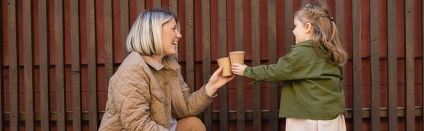 side view of cheerful woman and girl toasting with paper cups near wooden fence outdoors, banner - Photo, Image