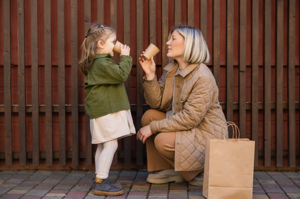 side view of mother and daughter drinking from paper cups near wooden fence and shopping bag - Photo, Image