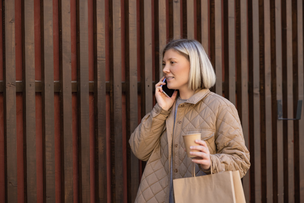 smiling blonde woman with coffee to go and shopping bag talking on smartphone near fence - Photo, Image