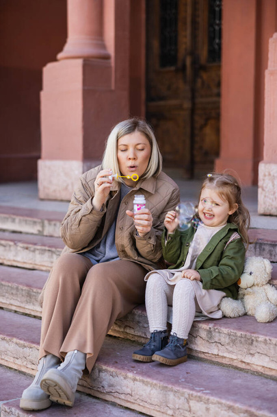 woman blowing soap bubbles near happy daughter on stairs outdoors - Φωτογραφία, εικόνα