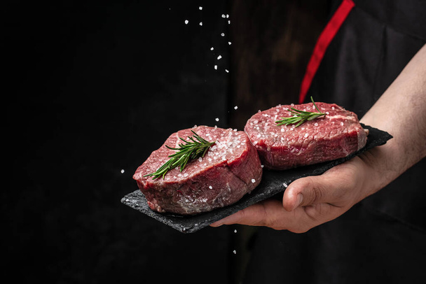Chef salts steak in a freeze motion with rosemary and spices. Preparing fresh beef or pork on a dark background. Long banner format. - Photo, Image