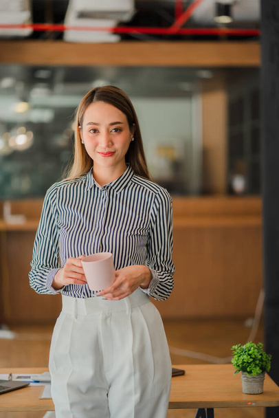 Happy successful Asian businesswoman holding a cup of coffee in front of the desk in the office - Fotó, kép