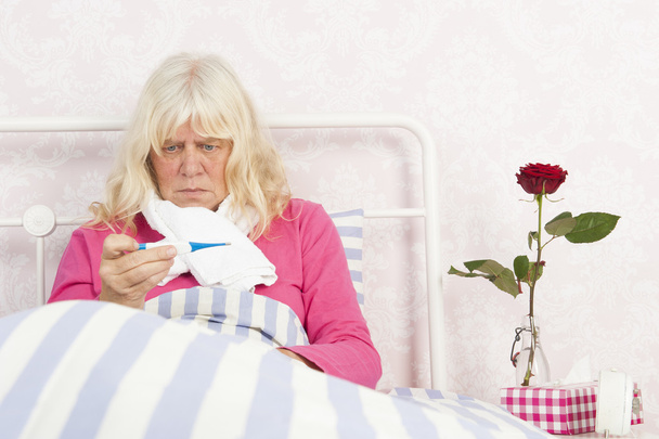 Γυναίκα που κάθεται στο κρεβάτι φιλοξενήσουμε με στο θερμόμετρο - Φωτογραφία, εικόνα