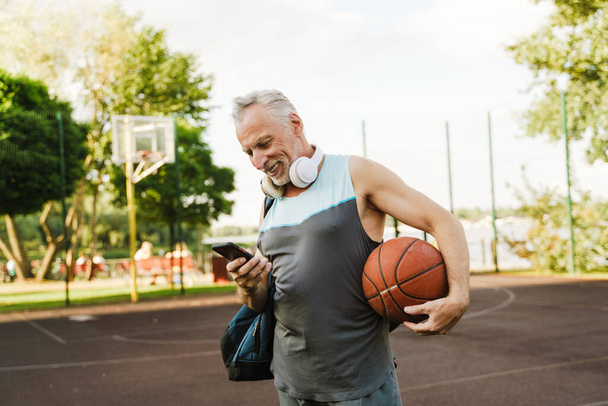 Grey senior man smiling and using cellphone on playground - Zdjęcie, obraz