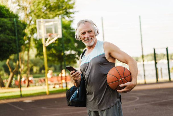 Grey senior man listening music and using cellphone on playground - Foto, afbeelding