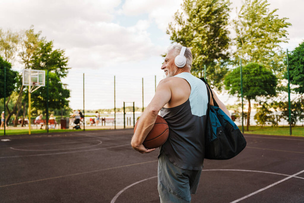 Grey senior sportsman smiling and listening music while working out on playground - 写真・画像