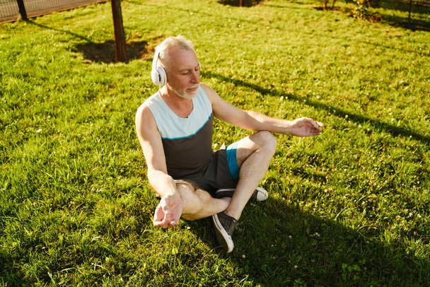 Senior man meditating and listening music during yoga practice in green park - Foto, afbeelding
