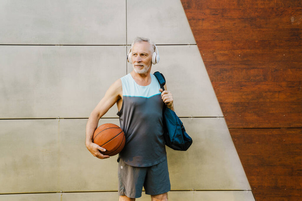 Grey senior man listening music and smiling while standing with ball by wall outdoors - Zdjęcie, obraz