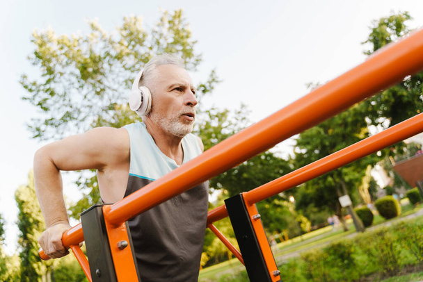 Grey senior man listening music while working out on equipment on sports ground - Foto, imagen