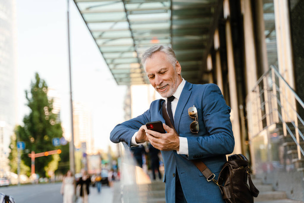 Senior man dressed in suit using mobile phone while standing by building at city street - 写真・画像