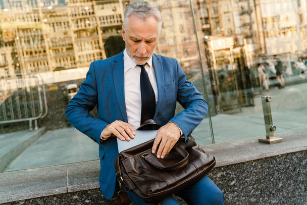 Senior man dressed in suit working with laptop while sitting by building at city street - Фото, изображение