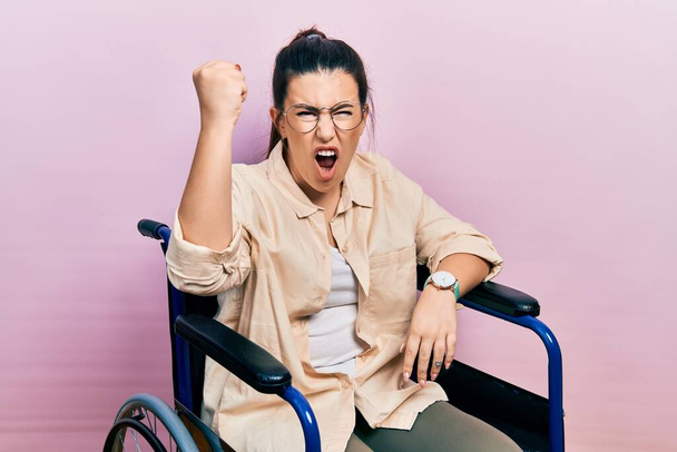 Tekerlekli sandalyede oturan genç İspanyol kadın kızgın ve kızgın yumruğunu kaldırıyor ve öfkeyle bağırıyor. öfke ve agresif konsept.  - Fotoğraf, Görsel
