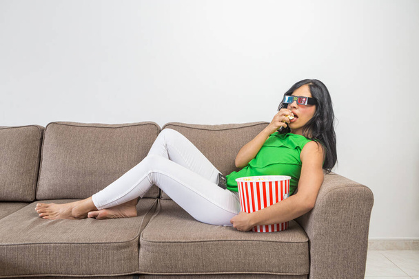 Side view of barefoot Hispanic female in casual clothes and 3D glasses eating fresh popcorn and watching interesting movie while chilling on couch in light living room - 写真・画像