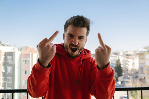 Young handsome man wearing red hoodie showing middle finger doing fuck you bad expression, provocation and rude attitude. Screaming excited. - Фото, изображение