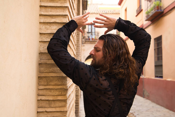 Long haired man dancing flamenco with black shirt and red roses. He makes dancing postures with his hands in a typical narrow street of Seville. Flamenco dance concept cultural heritage of humanity. - Photo, Image
