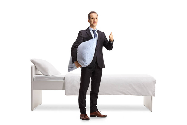 Businessman holding a pillow and showing thumbs up next to a bed isolated on white background - Photo, Image