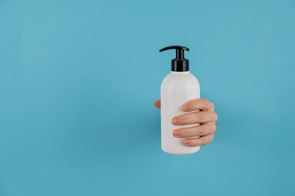 A womans hand sticks out of a hole in a blue paper background and holds a skin lotion pump. - Photo, Image