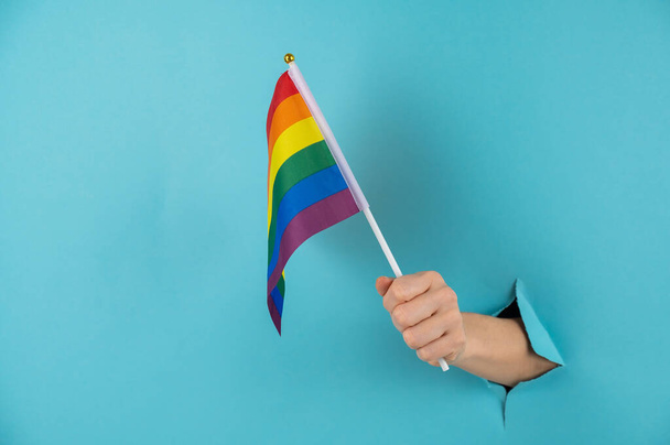 A womans hand sticks out of a hole in a blue paper background and holds an lgbt flag. - Foto, Imagem