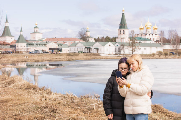 Mother and daughter take a family selfie against the backdrop of an ancient monastery and a spring landscape with a blue lake - Fotografie, Obrázek