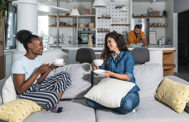 Two young woman sitting on the sofa drinking tea or coffee talking about female problems while man standing in the kitchen and cooking lunch for them reading recipe from the internet on laptop - Foto, Imagen