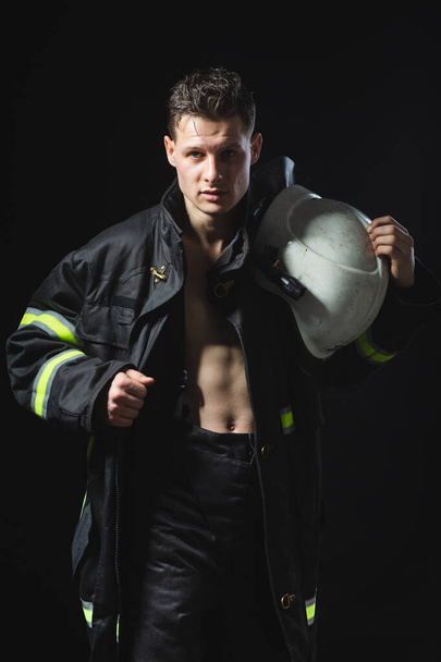 Módní portrét mladého muže v hasičském vybavení na černém pozadí studia. Hasič v tmavé uniformě - Fotografie, Obrázek